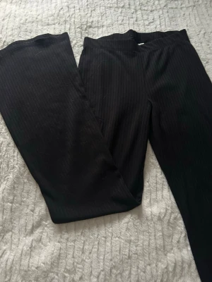 Svarta ribbade bootcut byxor - Säljer ett par svarta bootcut byxor med ribbad struktur. Byxorna har en mjuk och stretchig känsla, perfekt för dig som gillar bekväma och snygga plagg. De har en enkel design utan synliga detaljer och är helsvarta med diskreta vertikala ränder.