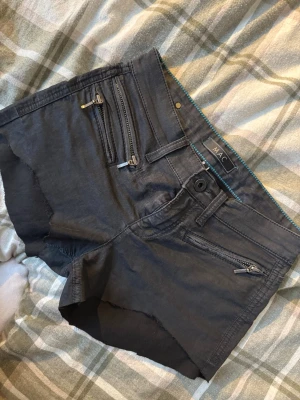 Svarta shorts från MAC Jeans med dragkedjor - Svarta avklippta shorts från MAC Jeans med flera snygga dragkedjor framtill. Modellen är lowwaist och har klassiska fickor både fram och bak. Materialet är en mix av bomull, polyester och elastan för skön stretch. Perfekt för dig som gillar en lite edgy stil. 36 cm rakt över. 