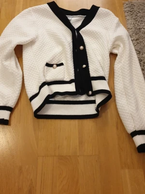 Vit och svart kofta från Lindex - Snygg vit kofta från Lindex med svarta detaljer och pärlknappar. Koftan har ett strukturerat stickat mönster, långärmade ärmar och en liten ficka framtill med pärldetalj. Passar dig som gillar klassisk och trendig stil.