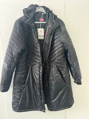 Svart lång pufferjacka från Áhkká- helt ny med lappar - Lång svart oanvänd pufferjacka från Áhkká med huva och quiltad design. Jackan har dragkedja framtill, praktiska fickor och är tillverkad i lätt syntetmaterial med värmande MóurráFill-fyllning. Perfekt för kalla dagar och riktigt snygg passform.