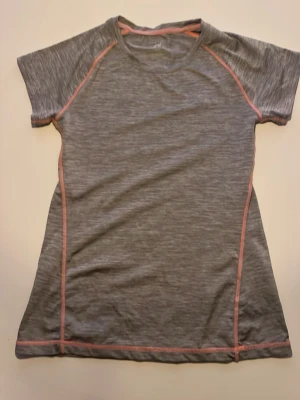 Grå sportig t-shirt med rosa sömmar - Säljer en grå t-shirt med korta ärmar och rosa kontrastsömmar. Perfekt för träning med sin sportiga look och lätt, syntetiskt material som andas. T-shirten har en rund hals och en normal passform som är bekväm att röra sig i.