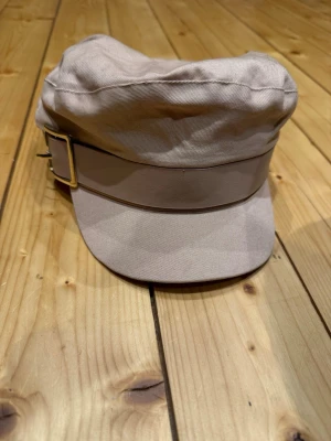 Laxrosa hatt - Laxrosa keps från River Island i en stilren och trendig modell. Kepsen har en kort skärm och ett dekorativt band med spänne framtill som ger en lite militär-/baker boy-inspirerad look. Perfekt accessoar för både vardag och outfitlyft.