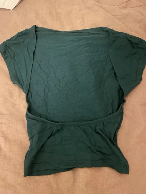 Mörkgrön croppad topp - Säljer en mörkgrön croppad topp med korta ärmar och öppen rygg. Toppen är tillverkad i mjuk bomull och har en enkel, stilren design utan mönster eller detaljer. Perfekt för dig som gillar minimalistisk stil och vill ha en bekväm topp till sommaren.