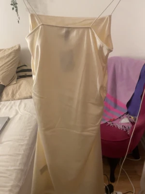 Champagnefärgad satinklänning S - Supersnygg champagnefärgad klänning i satin med tunna axelband och rak halsringning. Klänningen har en slät, glansig yta och är figurnära upptill med en lätt utställd kjol. Perfekt för dig som vill ha en elegant och stilren look.