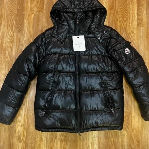 Svart pufferjacka från Moncler M - Säljer en svart pufferjacka från Moncler i storlek M. Jackan har huva, två dragkedjeförsedda fickor och klassisk Moncler-logga på ärmen. Materialet är glansigt och känns riktigt lyxigt, perfekt för kalla dagar. Stängs med dragkedja framtill.