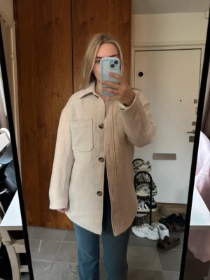 Beige overshirt med stora fickor - Snygg beige overshirt med stora bröstfickor och knäppning framtill. Jackan har en rak passform, klassisk krage och är tillverkad i ett mjukt material som känns lite som ull. Perfekt att slänga över en hoodie eller t-shirt för en chill look. Stor i storlek, passar mig som har storlek S/M