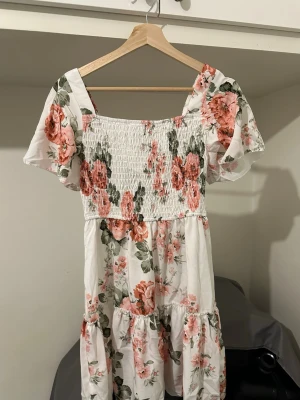 Blommig kortklänning från Elenzga - Supersöt kortklänning med vita och rosa blommor från Elenzga. Klänningen har smockad överdel, volangig kjol och korta ärmar. Perfekt för varma dagar och har en romantisk vibe. Materialet känns lätt och luftigt.