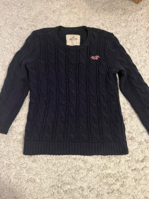 Mörkblå stickad tröja från Hollister - Säljer en mörkblå kabelstickad tröja från Hollister med rund hals och diskret logga på bröstet. Tröjan har korta armar så att de sitte fint! Använd några få gånger!💞