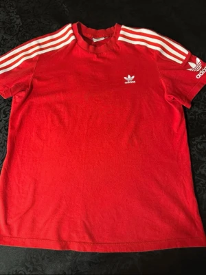 Röd Adidas t-shirt med vita ränder - Säljer en röd Adidas t-shirt med klassiska vita ränder längs axlarna och ärmarna. T-shirten har korta ärmar, rund hals och Adidas logga tryckt både på bröstet och ena ärmen. Skön bomullskvalitet som passar perfekt till streetwear och chill.