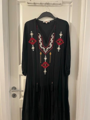 Svart långklänning med broderi från Koton - Svart långklänning från Koton med färgstarka broderade detaljer i rött, beige och gult framtill. Klänningen har v-ringad hals, trekvartsärmar och dekorativa tofsar. Materialet är mjukt och lätt, perfekt för dig som gillar bohemisk stil.