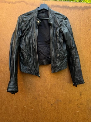 Svart skinnjacka från Rock'n Blue - Säljer en svart skinnjacka från Rock'n Blue med coola detaljer och dragkedjor. Jackan har en kortare passform, ståkrage med knapp och fickor med dragkedja. Perfekt för dig som vill ha en edgy look till vardags.