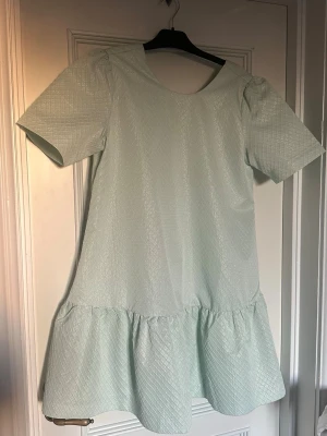 Ljusgrön kortklänning med struktur - Supersöt ljusgrön kortklänning med volang nertill och korta ärmar. Klänningen har ett diskret rutmönster i tyget som ger en glansig och lyxig känsla. Det är små guld detaljer i tyget som gör att det ser glittrigt ut. Perfekt för sommarens alla tillfällen! Materialet känns lite tjockare och har struktur.