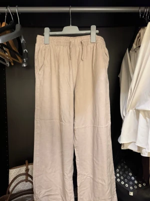 Beiga linnebyxor med snörning - Säljer ett par beiga linnebyxor med avslappnad passform och elastisk midja med snörning. Byxorna har raka ben, sidofickor och en bakficka. Perfekta för en chill och stilren look. Man kan även vika ner midjan och få dem Lågmidjade.