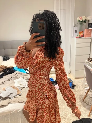 Röd och beige omlottklänning med mönster - Säljer en kort omlottklänning med röd och beige leopardmönster. Klänningen har långa puffärmar med smock vid ärmslut, v-ringad hals och volangdetaljer vid kjolen. Snygg knytning i midjan för extra form. Materialet känns lätt och följsamt.
