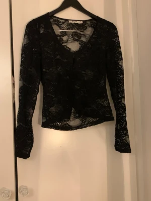 Svart spetskofta/blus från NA-KD - Säljer en svart kofta från NA-KD i  spets. Koftan har långa ärmar, knappar framtill och lös passform. 