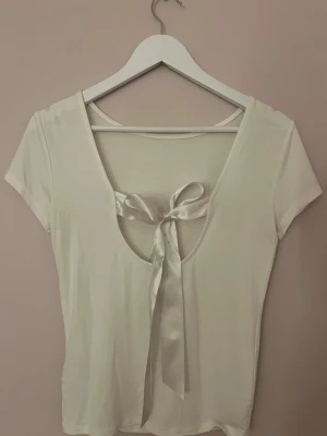 Vit topp med rosett från 157 - Supersöt vit topp från 157 med korta ärmar och en snygg öppen rygg som knyts med en rosett. Toppens material känns mjukt och stretchigt. Använd en gång.