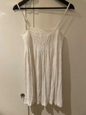 Vit somrig linneklänning med smock - Säljer en luftig vit linneklänning med smala axelband och smock upptill. Klänningen har en flowy passform och är gjord i ett tunt, mjukt material som passar perfekt till varma dagar. Enkel och stilren design som är lätt att matcha.