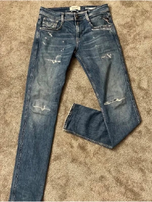 Replay Anbass blå jeans,  - Säljer ett par Replay Anbass jeans i klassisk blå tvätt med slitna detaljer och snygga slitningar på benen. Modellen har fem fickor, normal passform och raka ben. Jeansen är tillverkade i bomull och har Replay-logga på bakfickan samt läderpatch i midjan.
