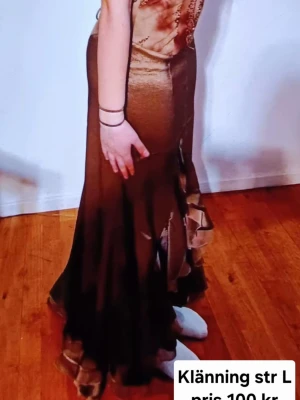 Brun långklänning med volang - Säljer en brun långklänning med snygga volangdetaljer nertill. Klänningen har en åtsittande passform upptill och faller snyggt längs kroppen. Materialet har en lätt glans och ger en elegant look. Perfekt för dig som vill sticka ut!