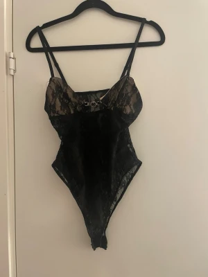 Svart spetsig korsettopp - Säljer en svart korsettbody i genomskinlig spets med tunna axelband och dekorativ detalj framtill. Bodyn är ifrån SHEIN och aldrig andvända pågrund av att den är förliten för mig, då jag är för lång för den.