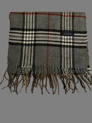 Grå rutig scarf från Burberrys - Säljer en klassisk grå rutig scarf från Burberrys i 100% lammull. Scarfen har ett tidlöst rutmönster i svart, vitt och röda detaljer samt fransar nedtill. Perfekt för dig som vill ha en ikonisk och stilren look. Perfekt nu när snön har kommit❄️ Kom med frågor eller prisförslag!