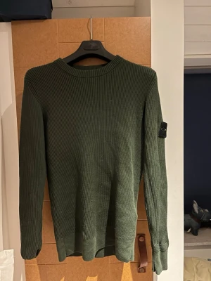 Grön ribbad stickad tröja Stone Island - Säljer en grön ribbad stickad tröja från Stone Island i storlek M. Tröjan har lång ärm och den klassiska Stone Island-loggan på vänster ärm. Perfekt för dig som vill ha en clean och stilren look med streetwear-vibe.