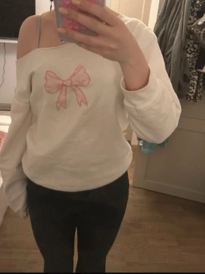 Offshoulder vit tröja med rosett - Säljer en vit offshoulder tröja med lång ärm och en stor rosa rosett broderad på bröstet. Tröjan har en avslappnad passform och är gjord i mjukt bomullsmaterial, perfekt för dig som gillar söta detaljer och comfy stil. Den är jättefin köp aldrig använd 🩷