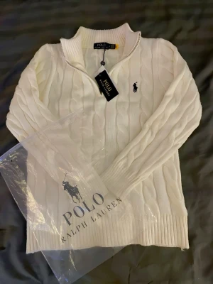Vit kabelstickad tröja Polo Ralph Lauren - Vit kabelstickad tröja från Polo Ralph Lauren i storlek S. Tröjan har half zip, ribbad krage och muddar samt den klassiska broderade loggan i svart på bröstet. Tillverkad i 100% bomull, perfekt för dig som gillar stilrena och tidlösa plagg.