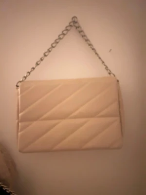 Beige quiltad axelväska med kedja - Säljer en beige axelväska med quiltad design och silverfärgad kedja som axelrem. Väskan har magnetlås och ett stort fack med innerficka med dragkedja. Perfekt storlek för det viktigaste och ger en clean look till outfiten.