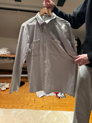 Ljusgrå skjorta från boohooMAN - Säljer en stilren ljusgrå skjorta från boohooMAN i storlek M. Skjortan har två stora bröstfickor, klassisk krage och knappar hela vägen framtill. Materialet är mjukt och känns som bomull, perfekt för en chill och clean look.