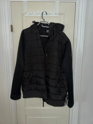 Svart pufferjacka från Ragwear - Svart pufferjacka med huva från Ragwear. Jackan har quiltad framsida och rygg, stickade ärmar och två dragkedjeförsedda fickor. Perfekt för kyliga dagar och har en modern, sportig look med ribbade muddar och hel dragkedja framtill. Den är som ny. 