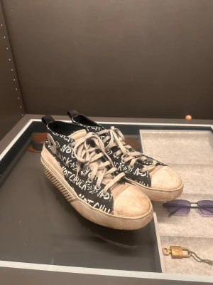 Converse Chuck Taylor - Unika svarta sneakers med vit graffiti-inspirerad text och beige detaljer. Skorna har snörning, rund tå och en kraftig platt sula i beige. Tillverkade i canvas med cool streetstyle-vibe och dragflikar bak för enkel påtagning.