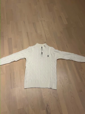 Vit kabelstickad tröja Polo Ralph Lauren - Kabelstickad vit tröja från Polo Ralph Lauren med half zip och klassisk logga på bröstet. Tröjan har lång ärm, ribbade muddar och hög krage. Perfekt för dig som gillar stilrena och tidlösa plagg.