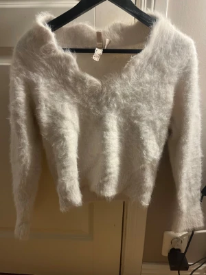 Vit fluffig stickad tröja DIVIDED XS - Supermjuk och fluffig vit stickad tröja från DIVIDED i storlek XS. Tröjan har lång ärm, v-ringad hals och en riktigt cozy look. Perfekt för dig som vill ha en mysig vibe till hösten eller vintern.