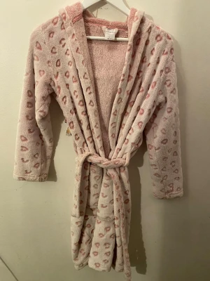 Rosa fleece morgonrock KappAhl hjärtan - Supermjuk och fluffig rosa morgonrock från KappAhl i storlek 146/152. Den har ett gulligt hjärtmönster i ljusrosa och bälte i midjan. Perfekt för mysiga hemmakvällar och har huva för extra komfort.