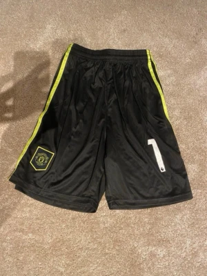 Svarta Manchester United shorts - Säljer ett par svarta Manchester United shorts med gula detaljer längs sidorna och klubbmärke på vänster ben. Siffran 1 i vitt på höger ben. Perfekta för fotboll eller träning.