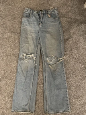 Blå ripped jeans från Vero Moda - Säljer ett par ljusblå jeans från Vero Moda med raka ben och stora slitningar över knäna. Jeansen har klassisk femficksdesign och är tillverkade i denim. Perfekta för en avslappnad och trendig look.