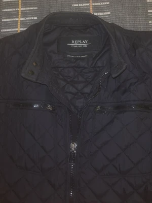 Svart quiltad jacka från Replay - Säljer en svart quiltad jacka från Replay med coola dragkedjor på bröstet och ärmarna. Jackan har ståkrage med tryckknappar och snyggt rutmönster över hela ytan. Perfekt för dig som gillar stilren och sportig look. Storlek M men passar även S 