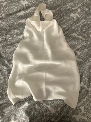 Vit halterneck linnetopp i satin - Säljer en stilren vit halterneck linnetopp i glansigt satinliknande material. Toppen har öppen rygg och knäpps i nacken. Perfekt för dig som gillar minimalistisk och chic stil. Passar till både jeans och kjol för en trendig look. Dubbeltyg över bysten så inte så genomskinlig