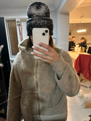 Beige teddyjacka med huva - Mysig beige teddyjacka med huva och dragkedja framtill. Jackan har två fickor och är perfekt för kyliga dagar. Den fluffiga ytan ger en trendig och avslappnad look.