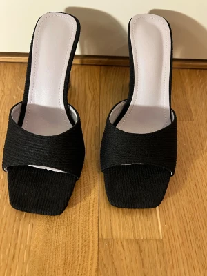 Svarta klackade sandaler med fyrkantig tå - Säljer ett par svarta sandaler med bred rem och fyrkantig tå. Skorna har en unik, genomskinlig blockklack och öppen tå. Ovandelen är i textil med struktur och innersulan är ljus. Perfekta för dig som vill sticka ut med en modern look.