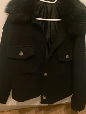 Svart teddyjacka med fuskpäls - Säljer en svart teddyjacka med stor krage i fuskpäls och snygga guldknappar. Jackan har två fickor framtill, lite kortare passform. Perfekt för dig som vill ha en mysig men ändå trendig look.