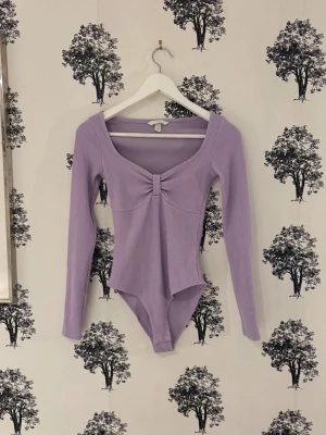 Ljuslila ribbad body med lång ärm - Supersnygg ljuslila body med lång ärm och ribbad struktur. Djup, rundad ringning framtill med twistad detalj vid bysten som ger en extra touch. Perfekt att styla med jeans eller kjol för en trendig look.
