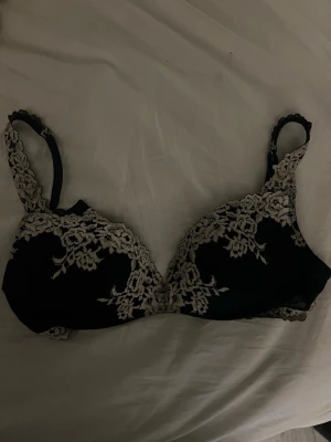 Svart spets push up bh INTIMISMI - Snygg svart bh med detaljerad blommig spets i vitt längs kupor och axelband. Justerbara axelband och dekorativ spetskant även baktill. Perfekt för dig som gillar en feminin och elegant look.