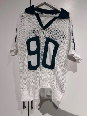 Vit mesh t-shirt med tryck Los Angeles - Säljer en vit mesh t-shirt med gröna detaljer och stort tryck med siffran 90 på framsidan. På ryggen står det Los Angeles i samma gröna färg. T-shirten har korta ärmar, ribbad kant och v-ringad krage med grönt parti. Materialet är stickad polyester med genomskinlig meshstruktur. 1XL but more L