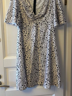 Vit blommig klänning med v-ringning - Superfin vit kort klänning med svartblommigt mönster och v-ringning. Klänningen har korta ärmar och ett lätt, flowy tyg. Snygg till festen eller ”style ner” den med ett par sneakers eller boots.