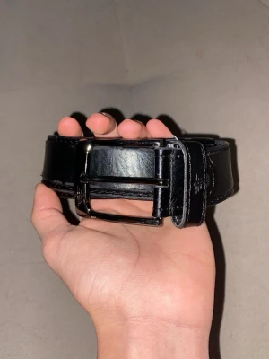 Svart Armani skärp i skinn med metallspänne - Säljer ett svart skärp i skinn med ett stilrent metallspänne. Skärpet har klassisk design med flera hål för justering och diskret logga vid spännet. Perfekt för att ge outfiten en clean och snygg touch.