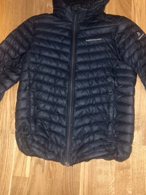 Svart Peak Performance - Säljer en svart dunjacka från Peak Performance med huva och dragkedja framtill. Jackan är quiltad med horisontella och sneda sömmar för en sportig look. Perfekt för kyliga dagar och enkel att matcha med det mesta.