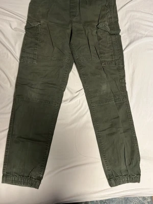Gröna cargopants från H&M - Säljer ett par gröna cargopants från H&M med elastiska muddar vid bensluten och stora fickor på sidorna. Byxorna har regular fit och är gjorda i mjuk bomull. Perfekt för dig som gillar streetstyle och praktiska detaljer.