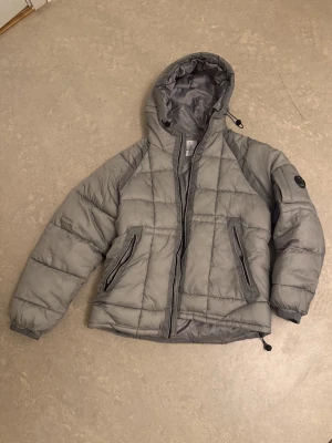 Grå pufferjacka från C.P. Company - Säljer en grå pufferjacka från C.P. Company med huva och dragkedja. Jackan har två snedställda fickor med dragkedja, justerbar dragsko i nederkant och en ikonisk linsdetalj på ärmen. Quiltad design och riktigt skön passform, perfekt för kalla dagar.